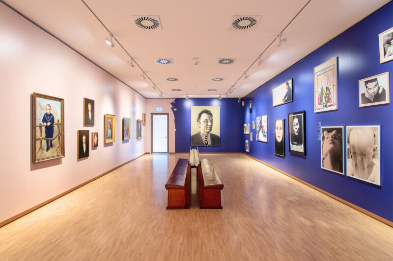 Gesicht ab! Ausstellungshalle Villa Mondriaan, Fotoporträts und gemalte Porträts hängen an weißen und blauen Wänden